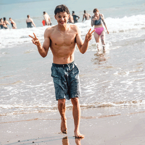 Person smiling and making peace signs while walking through shallow ocean water at a beach.